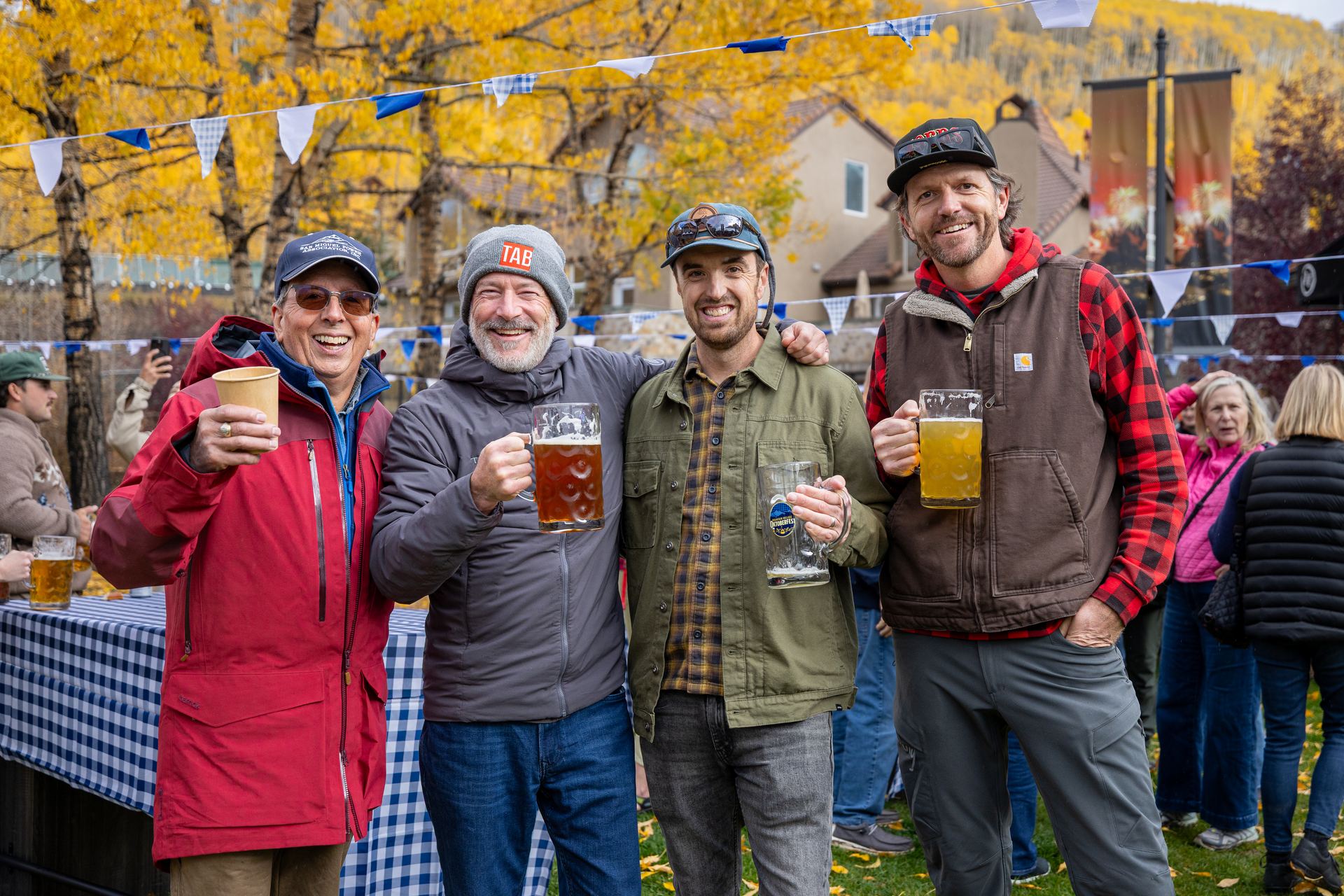 Four friends toasting beers outdoors under string bunting and golden aspens