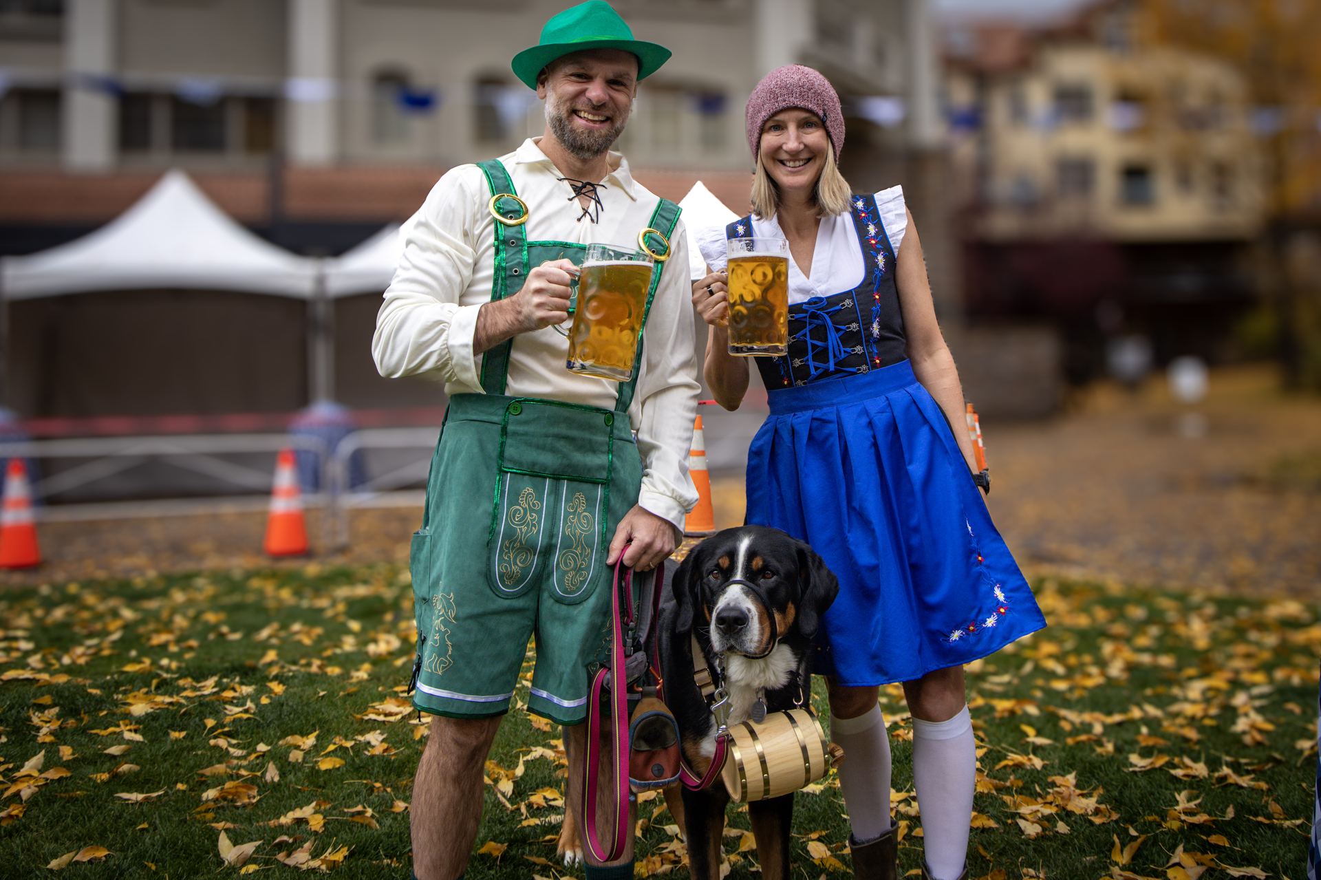 A couple in lederhosen and a dirndl toasting beers with their dog in a barrel collar